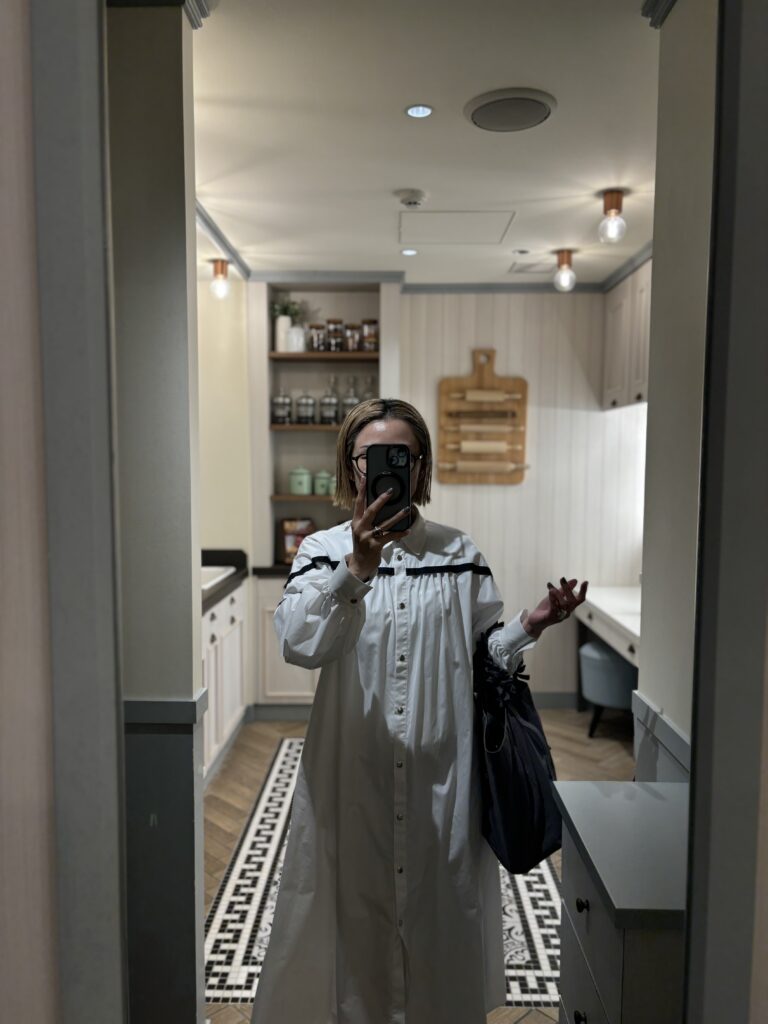 白いロングワンピースを着た女性が、室内のミラー越しに全身を自撮りしている写真。周囲には棚やライトがあり落ち着いた雰囲気。