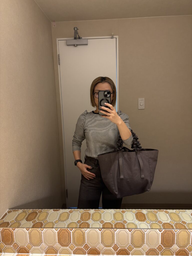 しまむらのトップスとGUのパンツを着た女性が、鏡の前でコーディネートを撮影している写真。大きめのグレーバッグを持っている。